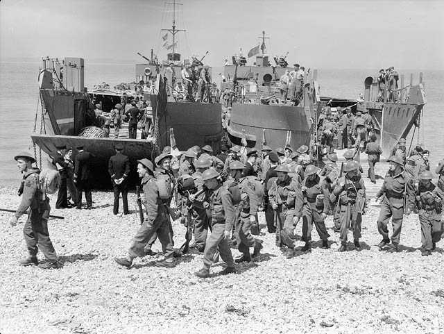 Black and white photograph. A large group of soldiers in battle dress walk onto a rocky beach from landing craft ramps. Navy personnel await the landing of a 2nd craft.